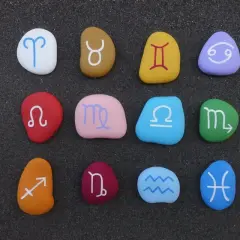Kolorowe kamienie z symbolami znaków zodiaku ułożone na ciemnym piasku.