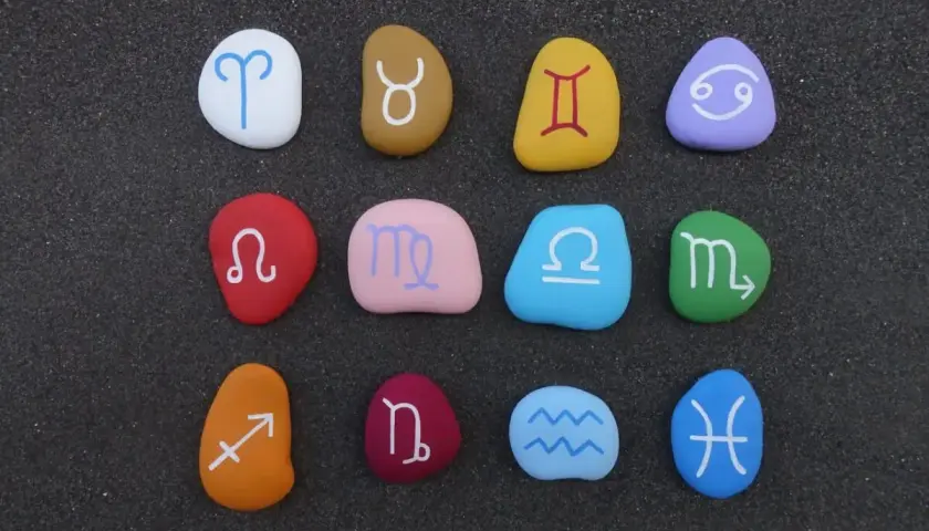 Kolorowe kamienie z symbolami znaków zodiaku ułożone na ciemnym piasku.