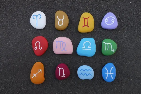 Kolorowe kamienie z symbolami znaków zodiaku ułożone na ciemnym piasku.
